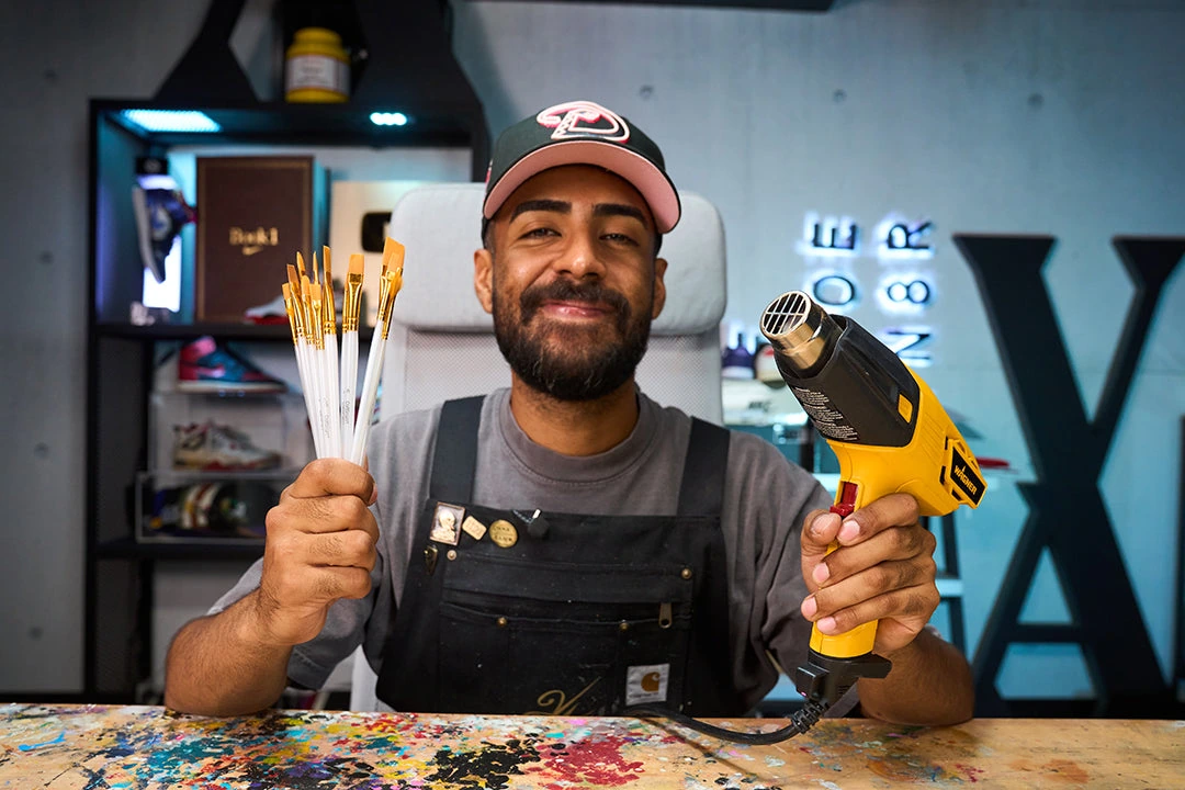 Vick Almighty smiling in a sneaker customization workshop, holding paint brushes and a heat gun